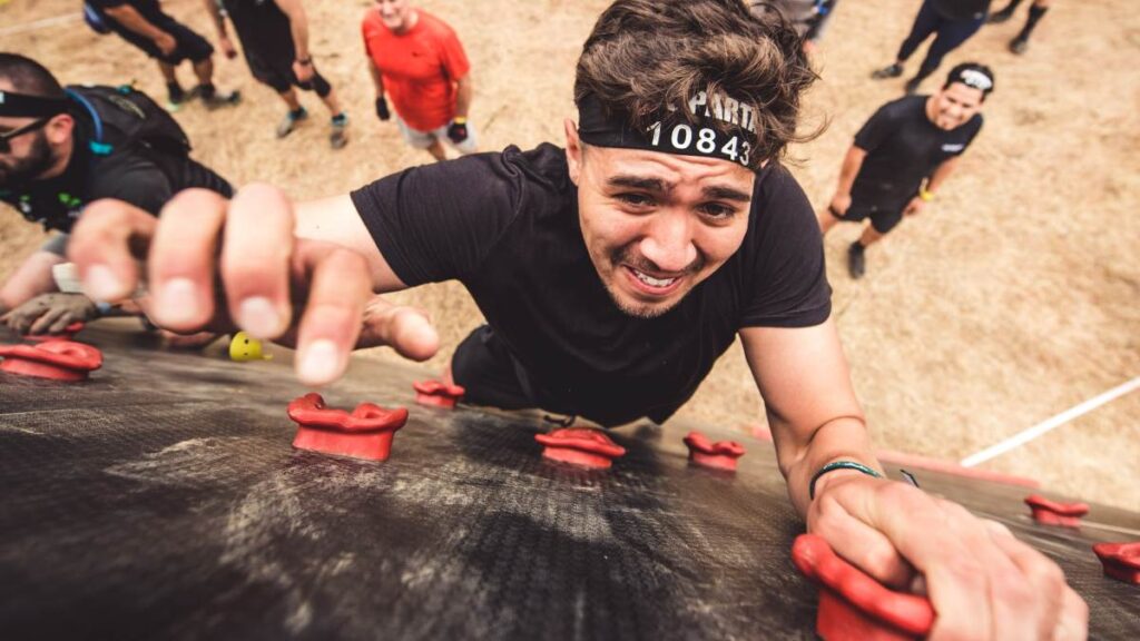 Hombre escala pared durante "Spartan Race" mientras otros lo observan.