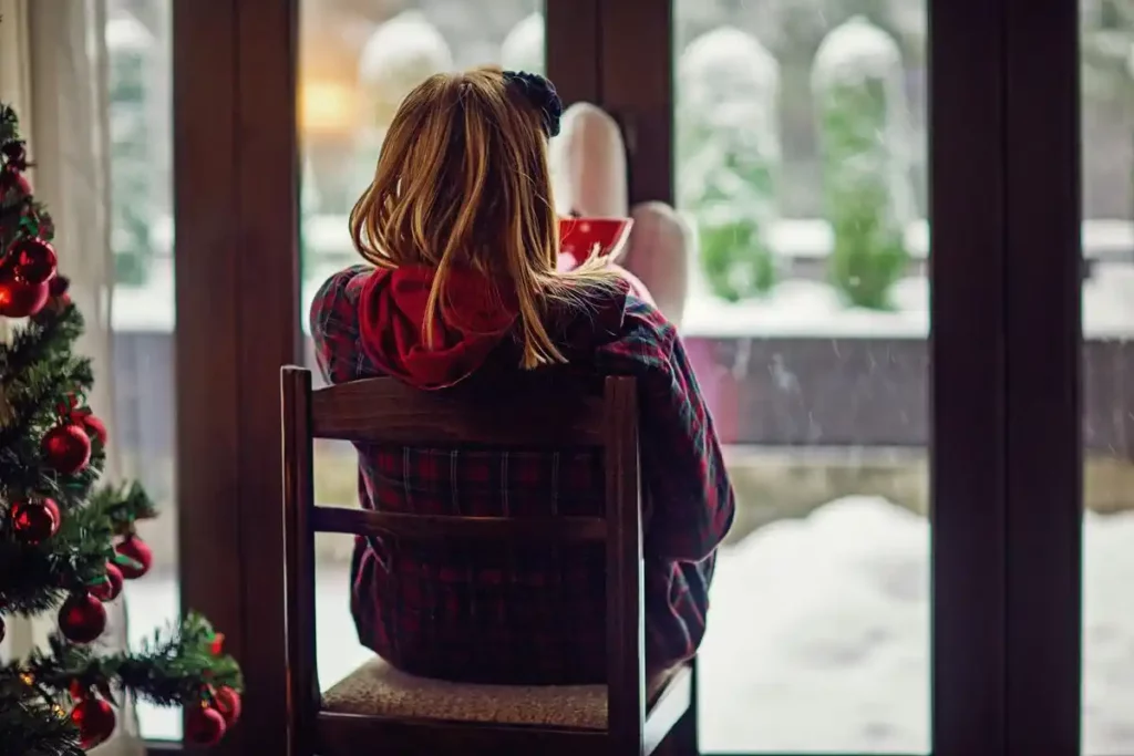 Mujer con depresión navideña sentada en una silla contemplando la nieve a través de una ventana.