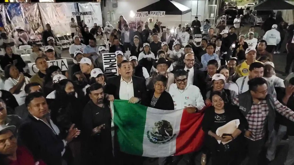 Alejandro Armenta con la bandera de México y migrantes poblanos en Estados Unidos.